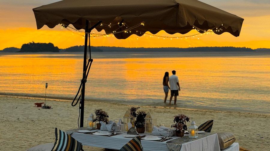 A shaded cabana setup on the beach with floor cushions and low tables, with 2 people standing near the shoreline at Silver Sand Sea Princess Beach Resort, Port Blair.